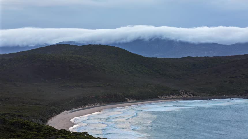 Coastal bay with a curved sandy beach rough sea and vegetationcovered hills under low clouds