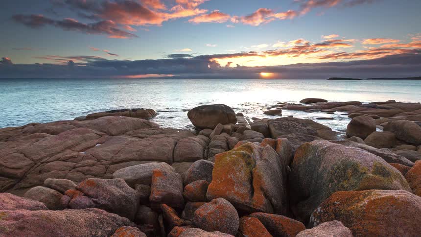 A rocky coastal shoreline at sunset with rugged rocks leading toward a calm ocean and glowing horizon