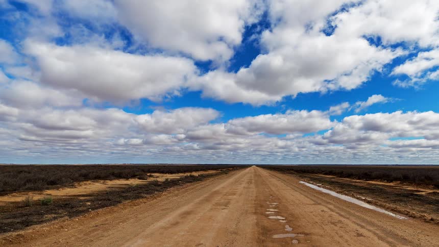A long straight dirt road stretches across a flat open landscape toward the horizon beneath a wide sky filled with scattered clouds