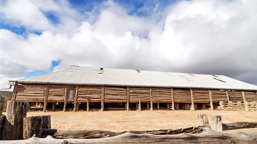 A long weathered wooden barn with a corrugated metal roof sits on open ground beneath a bright sky filled with drifting clouds