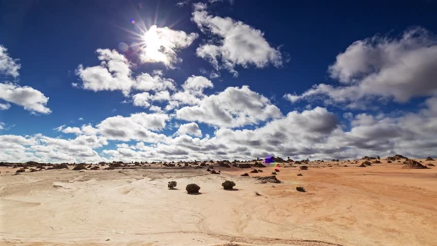 A vast desert plain with scattered rocks and sparse shrubs under a bright blue sky with white clouds