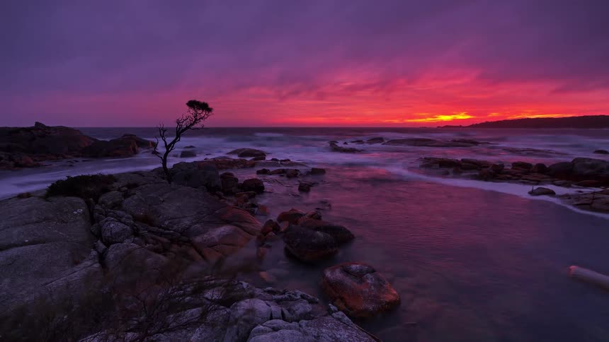 Water flows through rocks along the coast under a colorful cloudy sunset sky as a photographer moves his tripod