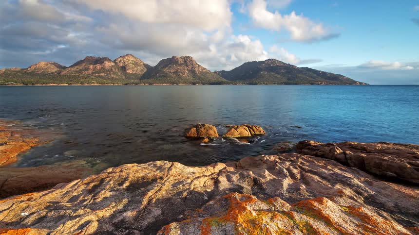 Jagged granite mountains rise above a calm blue bay under a cloudy sky and the foreground shows a rocky shoreline covered in patches of orange lichen while small waves gently wash over two central rocks sitting in the dark water of the coast