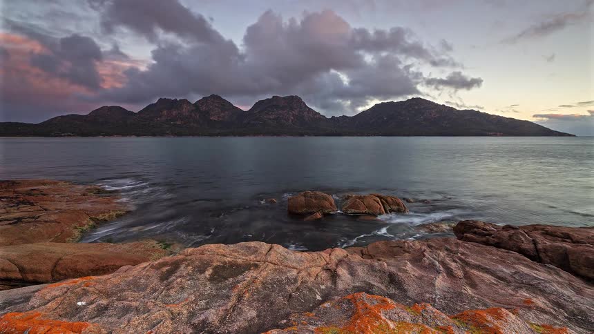 Clouds drift over jagged mountains and a rocky coastline as a golden sunset transitions into a purple twilight