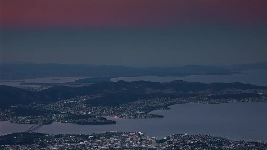 Aerial view of a coastal urban area with bays and mountainous terrain under a darkening sky at dusk