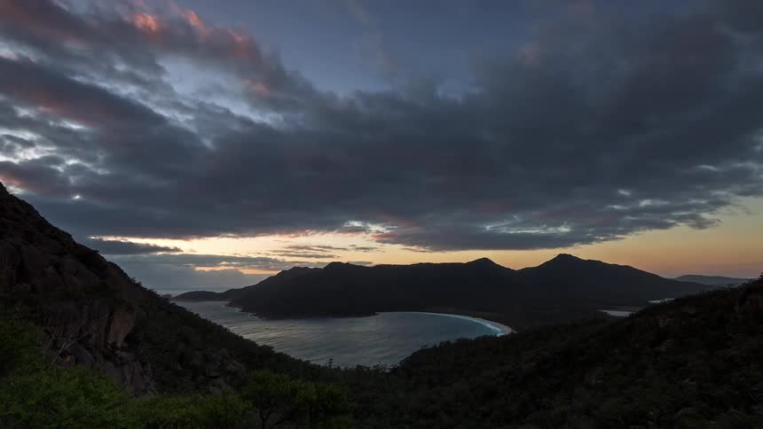 A high angle view shows a crescent shaped beach tucked between dark forested mountains and the ocean in a time lapse scene