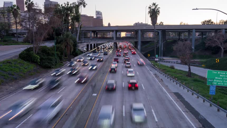 Rushhour traffic streams along a Los Angeles urban freeway at dusk with cars blurring past beneath overpasses and palm trees