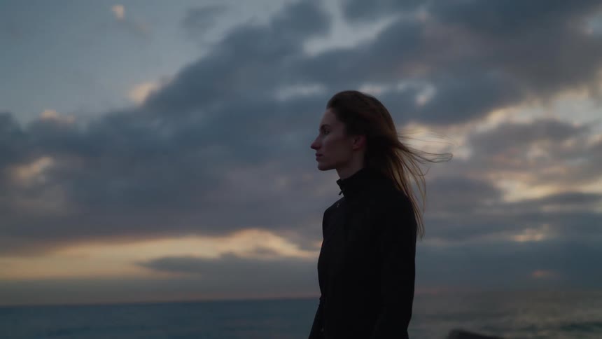 Person standing by the sea wearing dark clothing beneath a cloudy evening sky