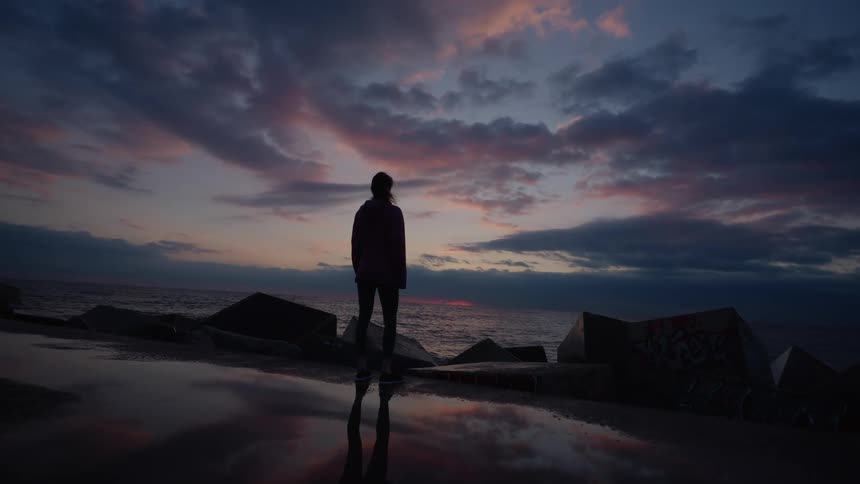 A person wearing a hoodie standing on a coastal road watching the ocean and rocky shoreline on a cludy evening