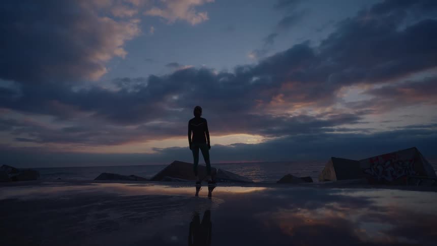 Silhouetted person standing on a wet surface by the sea with rock formations and a cloudy sky at dusk