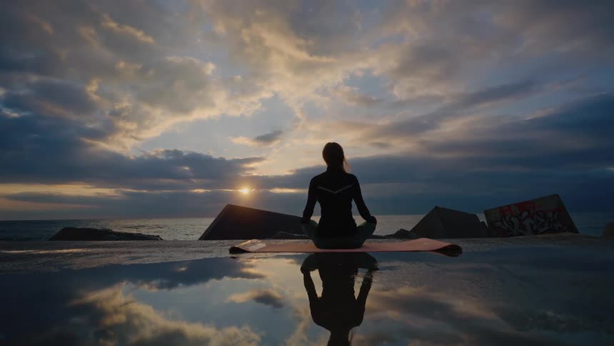 Person seated in a meditation posture by the sea with rocks reflection on a wet surface and a cloudy sky at dusk
