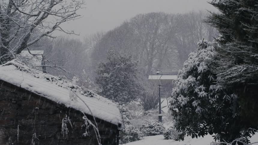 Heavy snow falls over a quiet winter garden where a stone building and a lamppost stand covered in white