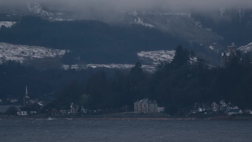 A wide shot shows a small coastal town nestled at the base of dark snowy mountains under a thick fog