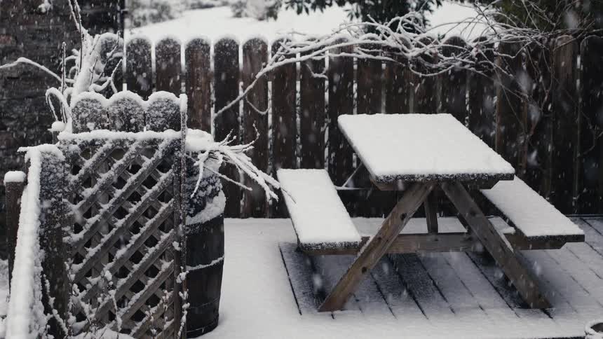 Heavy snow falls over a wooden picnic table and bench set sitting on a backyard deck