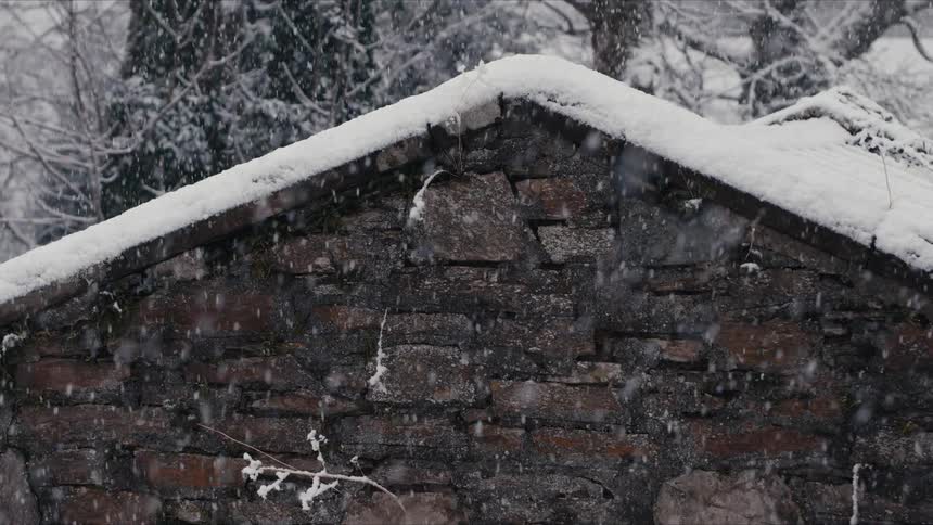 A rustic stone building with a snow covered gabled roof stands during a heavy snowfall as white flakes fall against the dark masonry and bare winter trees in the blurred background