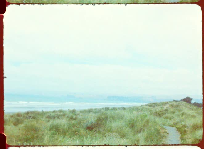 A narrow dirt path winds through green grassy dunes leading toward a wide sandy beach and a calm ocean under a vast overcast sky with distant cliffs visible through the haze