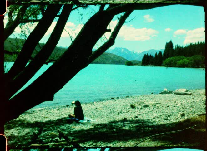 A woman sits on a rocky beach by a turquoise lake while large tree branches frame the view of distant mountains and a pine forest under a bright sky
