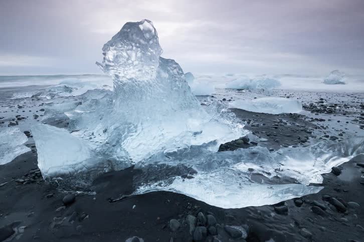 A massive translucent iceberg sits on a black sand beach covered in dark volcanic pebbles while smaller ice fragments float in the misty ocean under a heavy grey sky