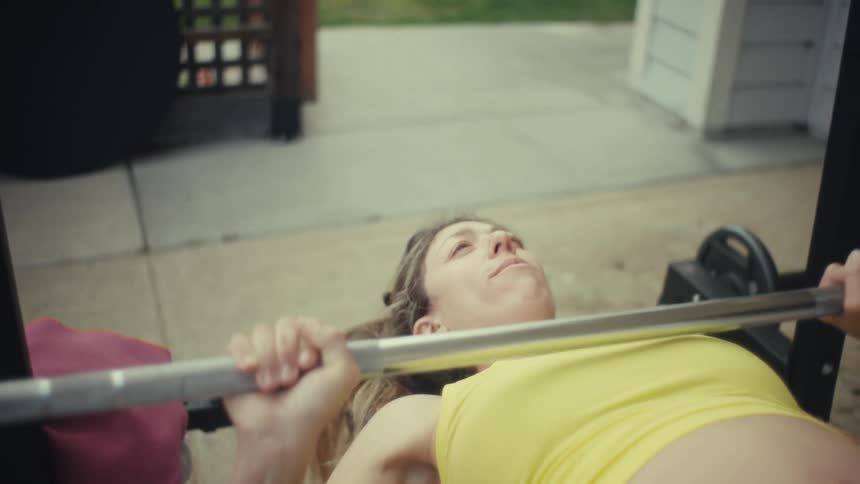 A young pregnant woman wearing a yellow singlet with a visible baby bump doing bench press weightlifting in an outdoor gym at midday