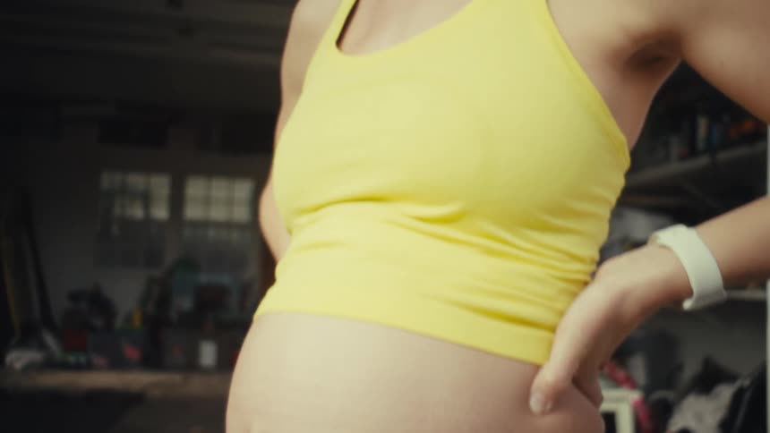 A pregnant woman in a yellow top and blue leggings stands in a garage holding her lower back while her bare stomach is shown in profile