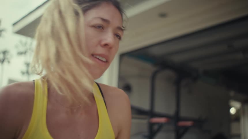 A pregnant woman with her hair in a ponytail wears a yellow tanktop while performing a physical exercise in a garage gym showing a focused expression of physical effort during her prenatal fitness routine as she moves on an exercise bench