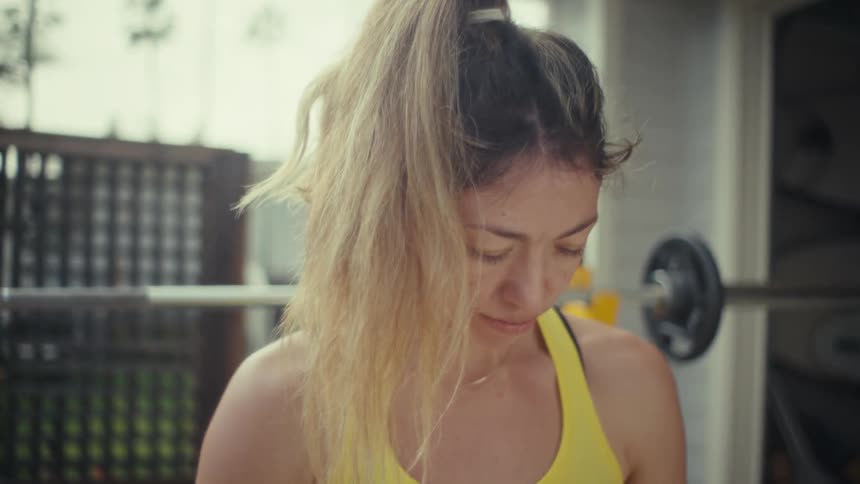 A pregnant woman wearing a yellow tanktop stands in an outdoor gym and holds her bare stomach while looking down near a barbell rack and a wooden fence during a daytime fitness session
