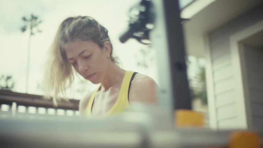 A young pregnant woman wearing a yellow tank top load weight plates onto a metal bar on a garage gym in a white residential building