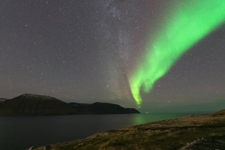 Northern lights over a coastal landscape with mountains in the background beneath a starfilled sky