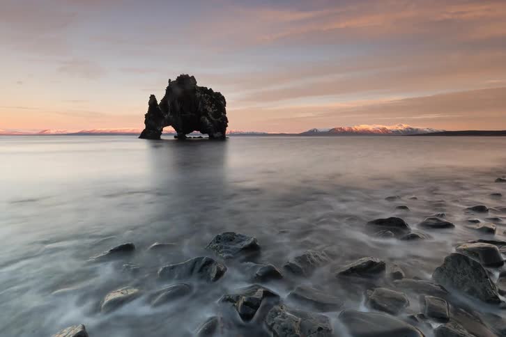 Isolated rock formation rising from calm water with dark stones in the foreground at dusk