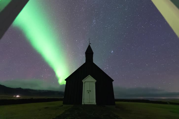 Arch in front of the black church in iceland as the green lights of the aurora borealis glow among stars in the sky