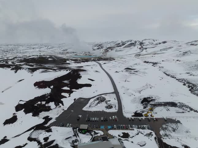 An aerial view reveals steam rising from a geothermal facility nestled within a vast snow covered volcanic landscape