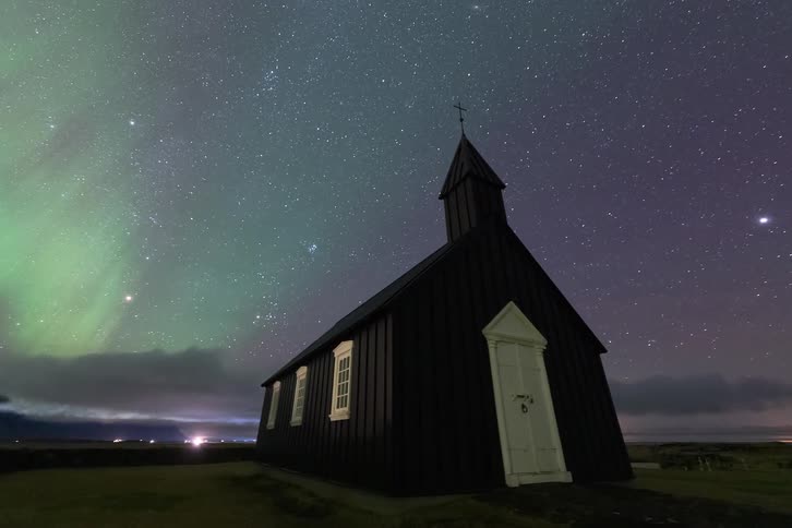 Wooden church on a field next to a road with traffic on it under the aurora borealis outdoors during night time