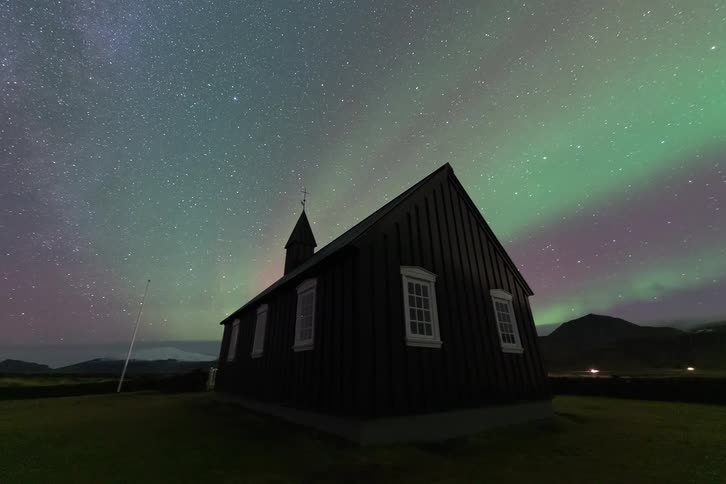 A time lapse view shows the vibrant green and purple aurora borealis and a field of stars moving across the night sky behind the silhouette of a small black church in a rural landscape