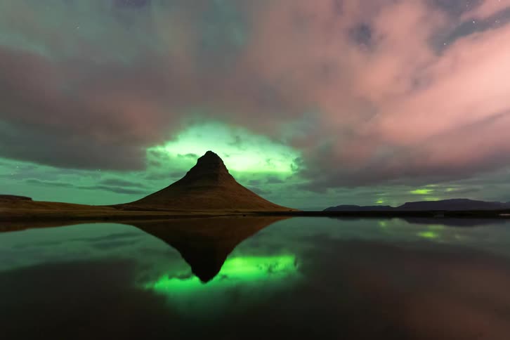 Aurora borealis reflected on a calm lake with an isolated mountain at the center under a night sky
