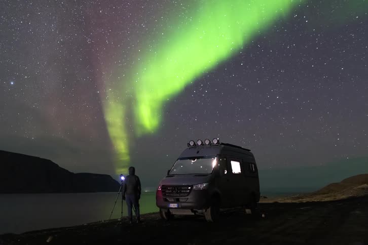 Northern lights in the night sky over a coastal area with a person photographing beside a parked vehicle