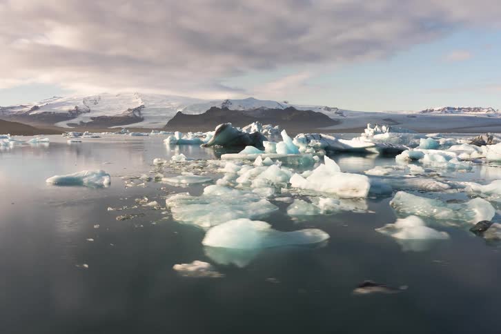 Icebergs swirl across a glassy polar lagoon their pale blue forms reflecting the cold light beneath distant snowcovered mountains and a brooding sky