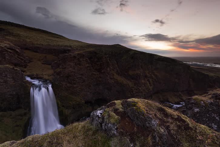 Waterfall in a rocky valley with surrounding hills beneath an evening sky