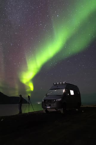 Photographer with his camera and tripod next to a van along the coast as the green lights of the aurora borealis glow over mountains in a starry sky