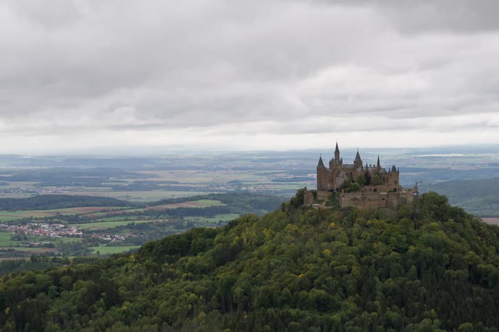 Historic castle atop a wooded hill with an expansive rural landscape in the background under a cloudy sky