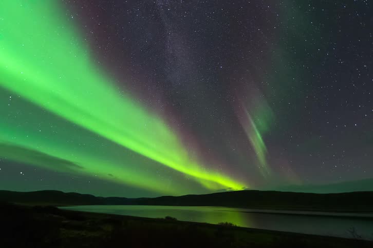 Vibrant band of neon green that originates from behind the dark silhouette of the hills on the left it sweeps upward and toward the center of the frame appearing as a solid concentrated beam of light