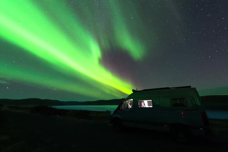 Intense northern lights over a coastal area with a parked vehicle and a person nearby beneath a starry sky