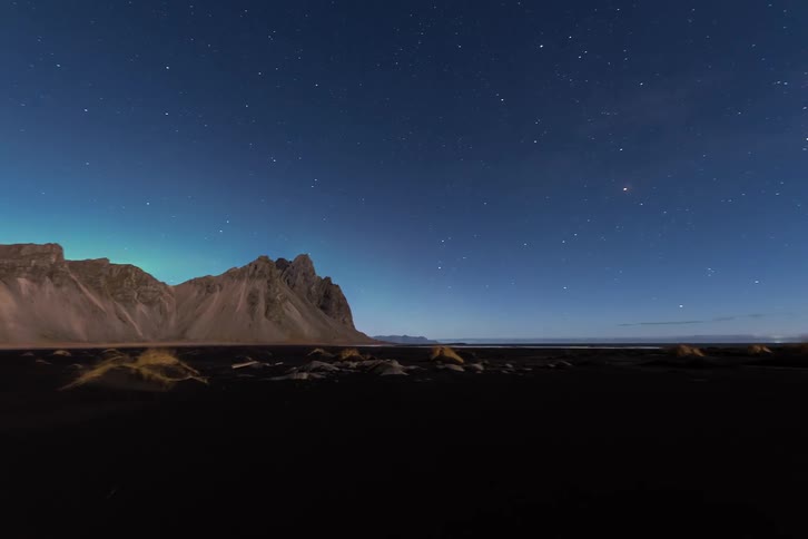 A light glows among rocks as the northern lights shine behind mountains under a starry sky