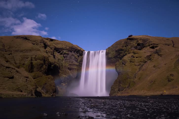 Waterfall illuminated at night between rocky cliffs with a visible rainbow in the mist