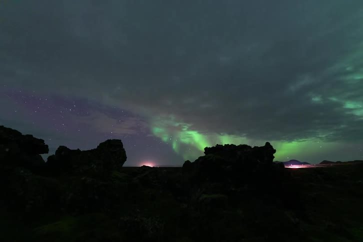 Northern lights visible on the horizon above rocky terrain beneath a cloudy night sky
