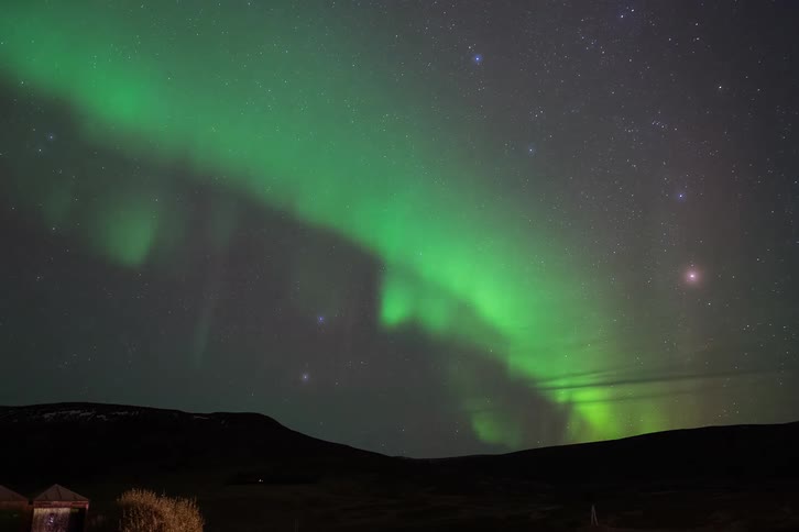 Northern lights forming a green arc above dark hills beneath a starry night sky