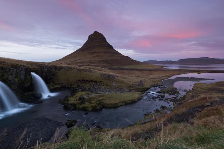 Hikers travel past a river and two waterfalls in hilly terrain as night falls
