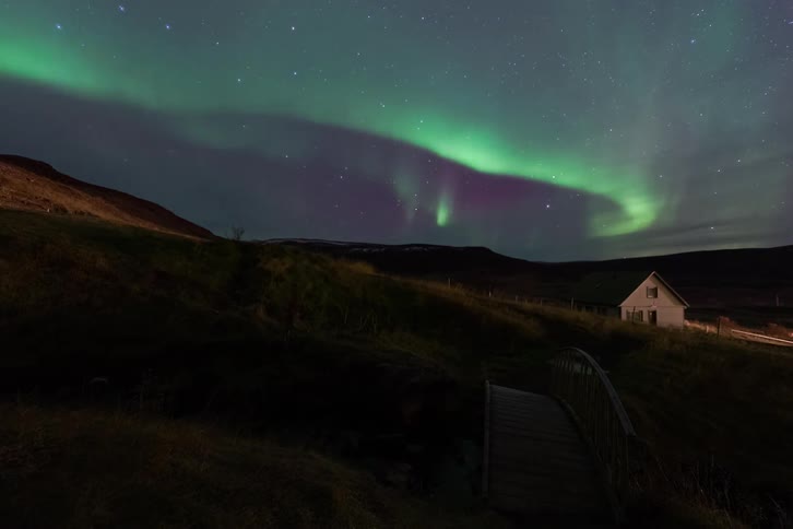 Aurora borealis arcing over a rural night landscape with an isolated house and a footbridge in the foreground
