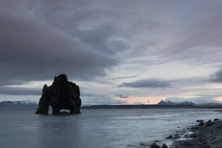 Isolated rock formation in the sea with a rocky shoreline and mountains in the background under a cloudy sky