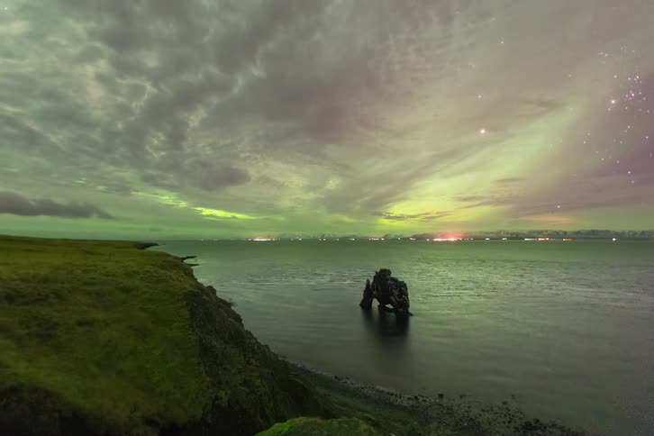 Aurora borealis over a rocky coastline with an isolated rock formation emerging from the water and a cloudy night sky