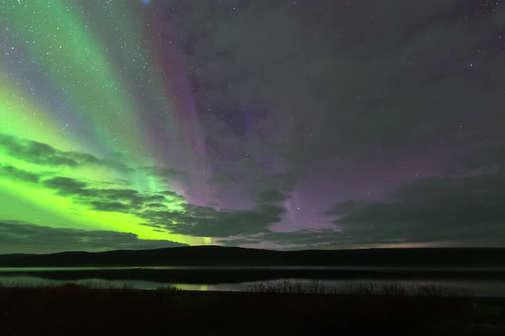 Northern lights coloring the night sky above a lake and silhouetted hills on the horizon